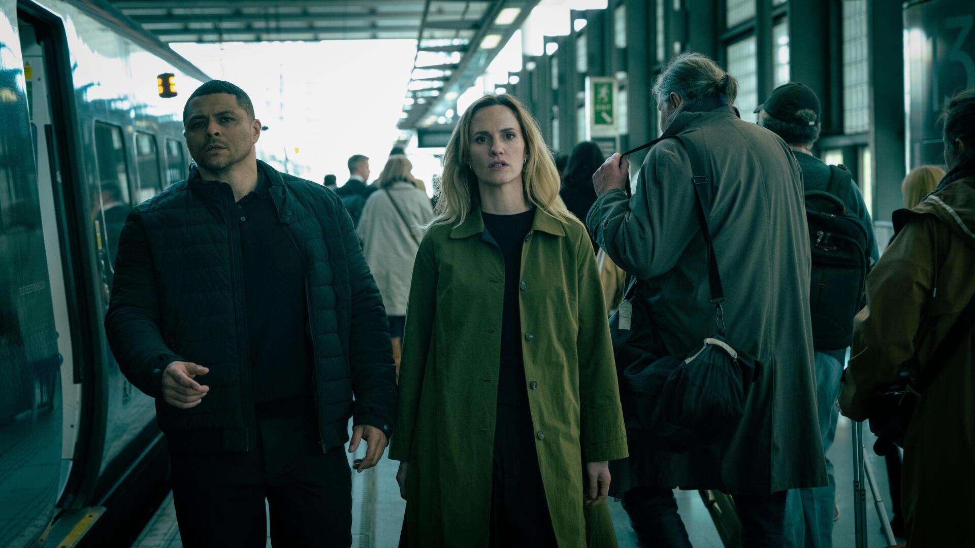 A scene from the TV show Slow Horses, featuring two characters walking through a busy train station. The image serves as an 8K Ultra HD desktop wallpaper and background.