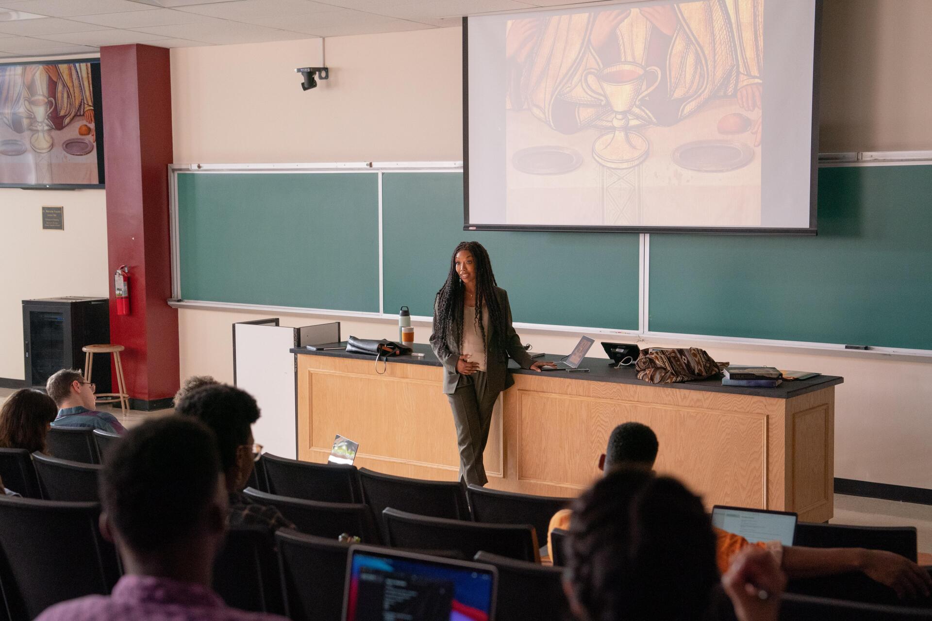A classroom scene from The Front Room, featuring Brandy Norwood engaged in a lecture, with students attentively listening. The setting includes a projector screen and multiple desks.