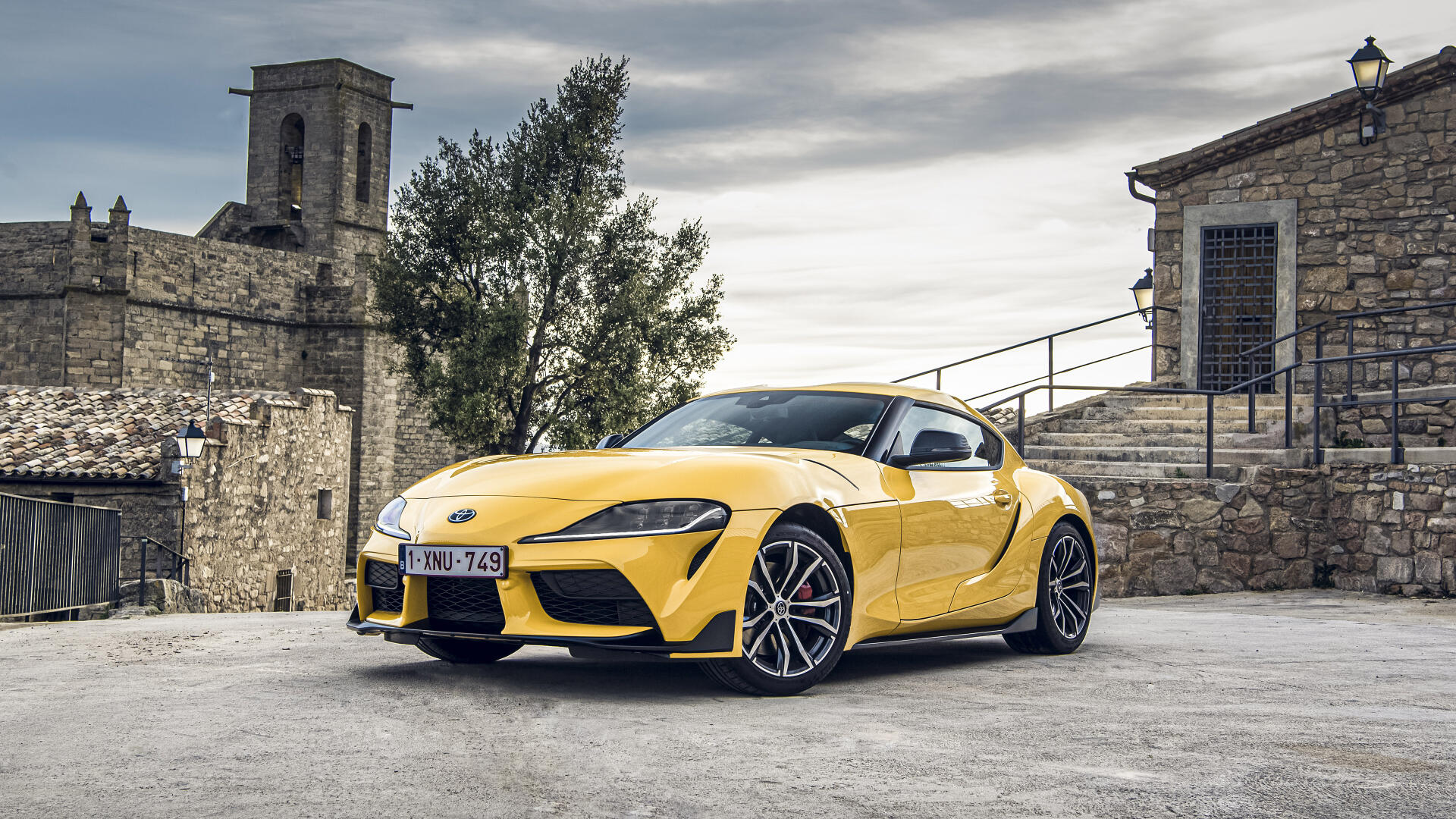 A striking yellow Toyota Supra parked in a scenic setting, showcasing its sleek design against a backdrop of rustic buildings and trees, captured in vibrant 4K Ultra HD.