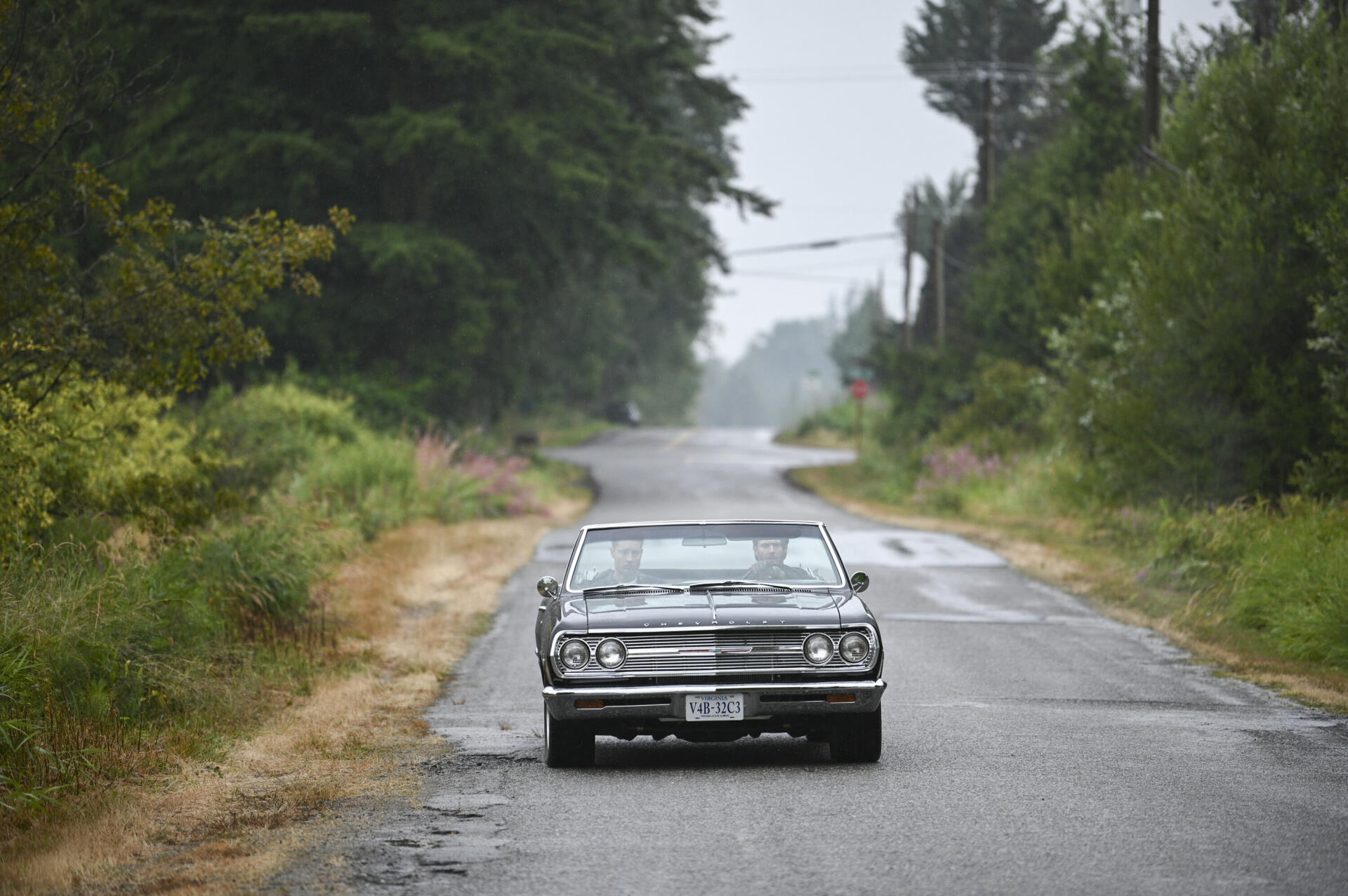 A classic car is parked on a quiet, rainy road, surrounded by lush greenery, serving as an atmospheric backdrop for the TV show Tracker (2024).