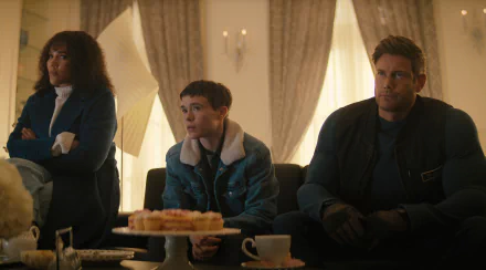 A scene from the TV show The Umbrella Academy, featuring three characters sitting on a couch with a plate of pastries in front of them, set in a warmly lit room.