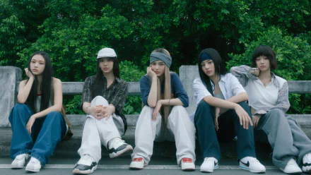 A stylish HD desktop wallpaper featuring the K-pop group NewJeans, showcasing five members sitting casually against a lush green background, embodying their unique aesthetic and music vibe.