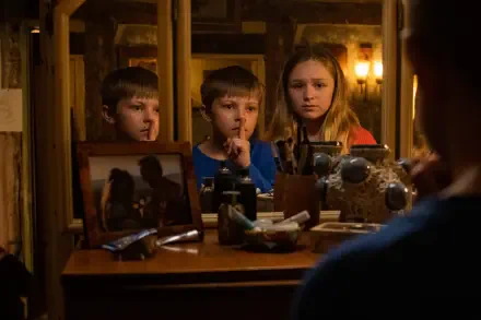 A tense scene from the movie Speak No Evil, featuring two children and a girl in front of a cluttered vanity mirror, whispering and sharing a secretive moment in warm lighting.