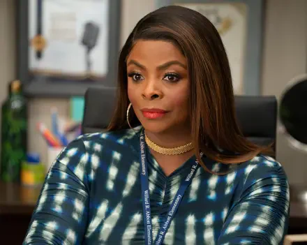 A close-up of a character from the TV show Abbott Elementary, showcasing a woman with long hair, wearing a patterned outfit and jewelry, seated in an office environment.