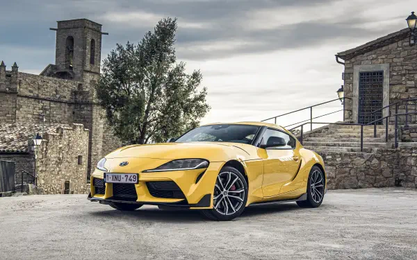 A striking yellow Toyota Supra parked in a scenic setting, showcasing its sleek design against a backdrop of rustic buildings and trees, captured in vibrant 4K Ultra HD.