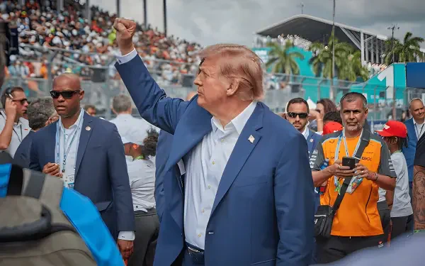 HD desktop wallpaper of a man in a navy suit raising his fist amid a crowded outdoor MAGA rally, flanked by security and supporters.