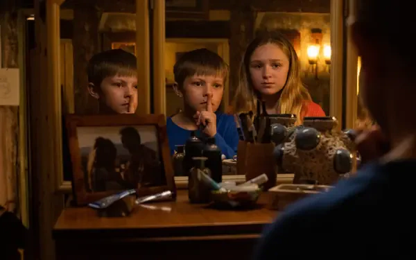 A tense scene from the movie Speak No Evil, featuring two children and a girl in front of a cluttered vanity mirror, whispering and sharing a secretive moment in warm lighting.
