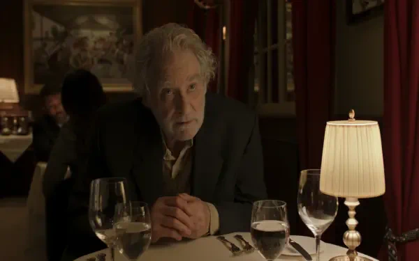 A dramatic scene from the TV show Disclaimer (2024) featuring Kevin Kline sitting at a restaurant table, with a soft-lit ambiance and wine glasses in view. 