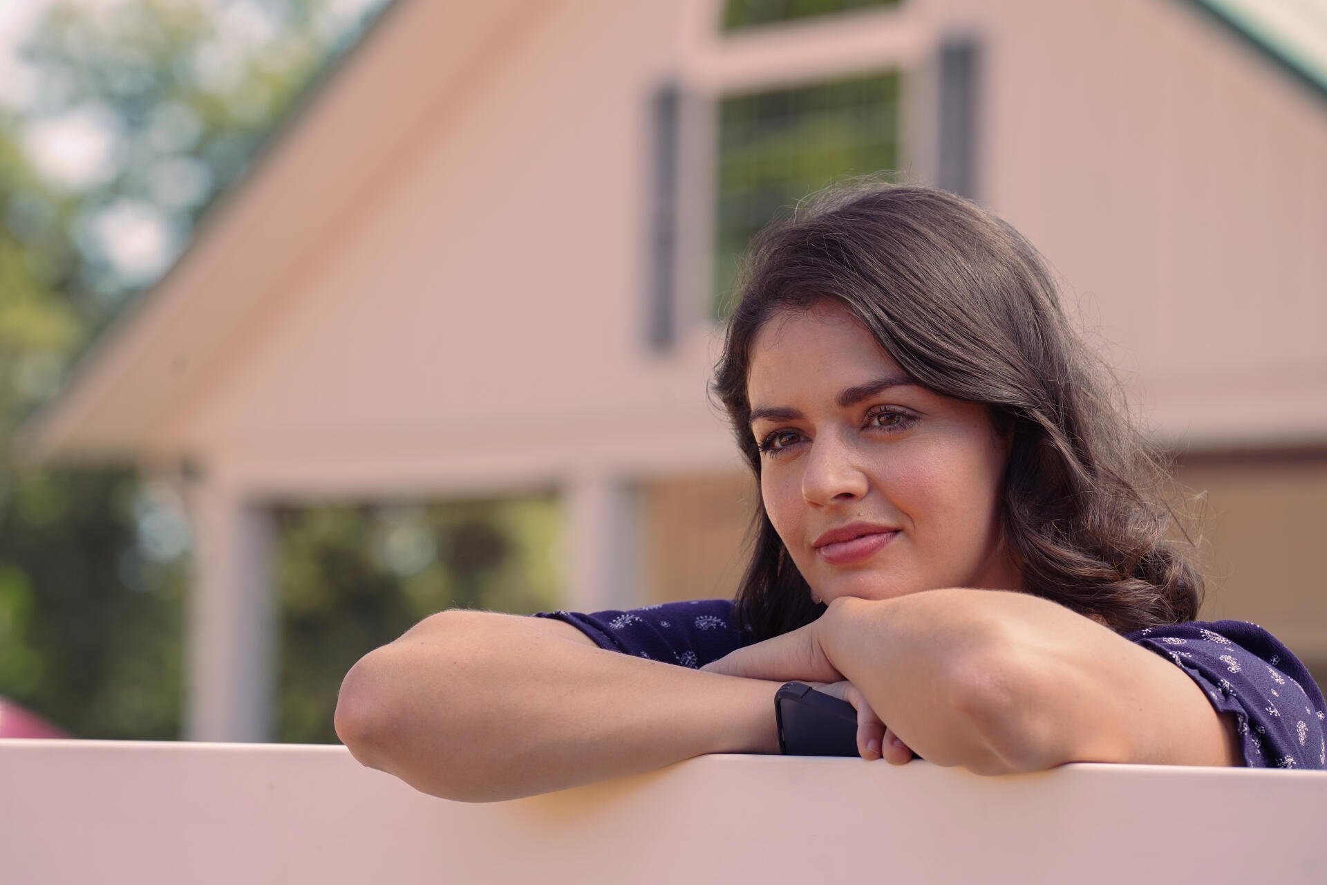 A woman leans on a white fence, gazing thoughtfully in front of a charming house, capturing a serene moment from the TV show Tulsa King, showcased as a 4K Ultra HD desktop wallpaper.