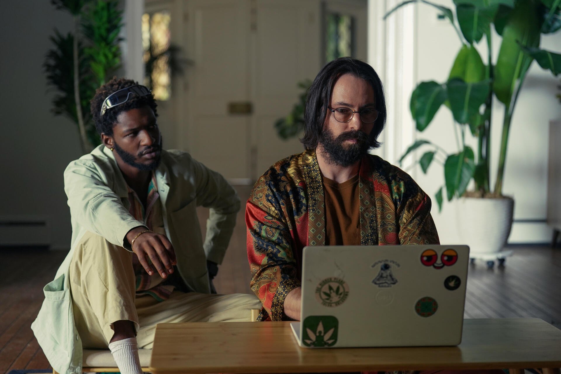 Two men are seated indoors, one working on a laptop while the other observes. The scene captures a moment from the TV show Tulsa King. 4K Ultra HD desktop wallpaper.