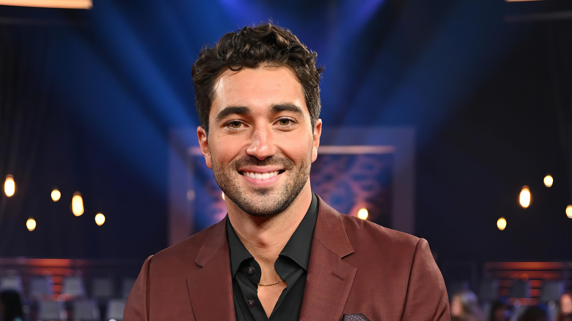 Joey Graziadei smiles confidently, dressed in a stylish suit against a backdrop of dramatic stage lighting, showcasing his role on Dancing with the Stars in this HD wallpaper.