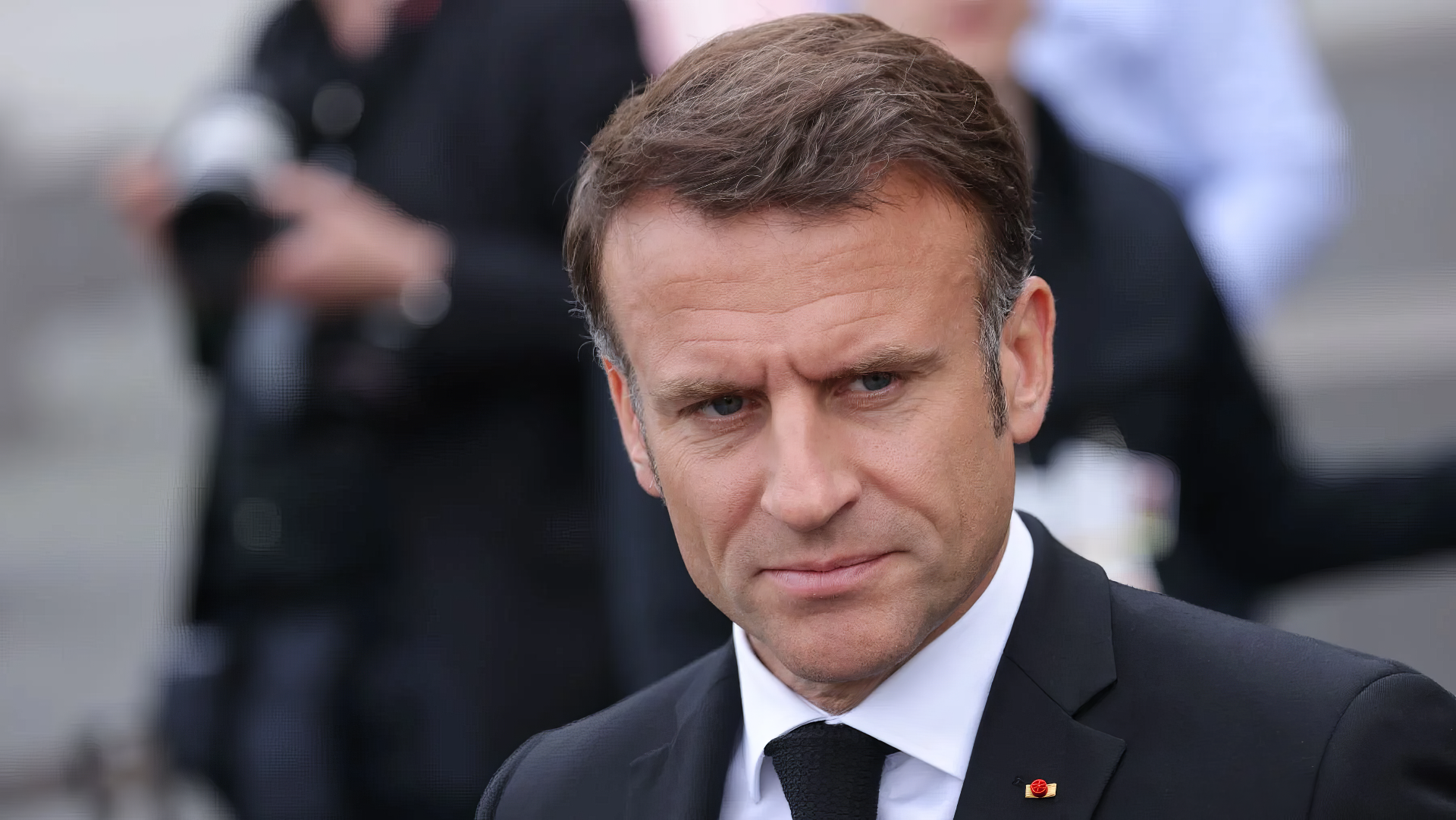 A close-up portrait of Emmanuel Macron, dressed in a black suit and tie, set against a blurred background of onlookers, designed as a HD desktop wallpaper and background.