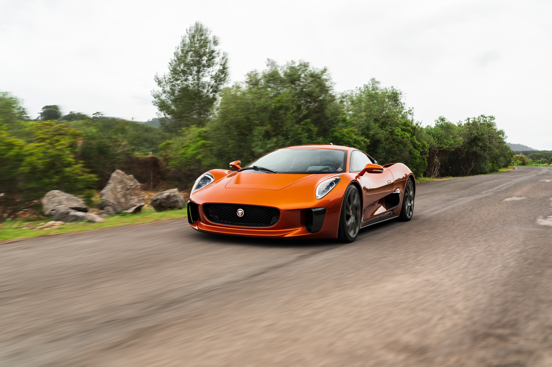 An orange Jaguar C-X75 sports car drives along a winding road, surrounded by lush greenery. This high-definition image serves as a striking desktop wallpaper and background.