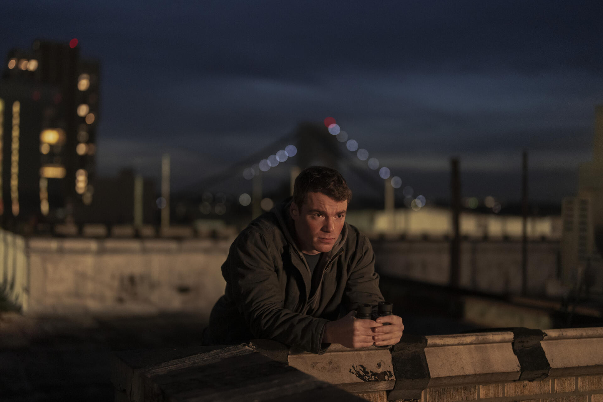 A dramatic scene from The Night Agent, featuring Gabriel Basso leaning against a rooftop barrier at night, with city lights and a bridge in the background. HD wallpaper-ready image.