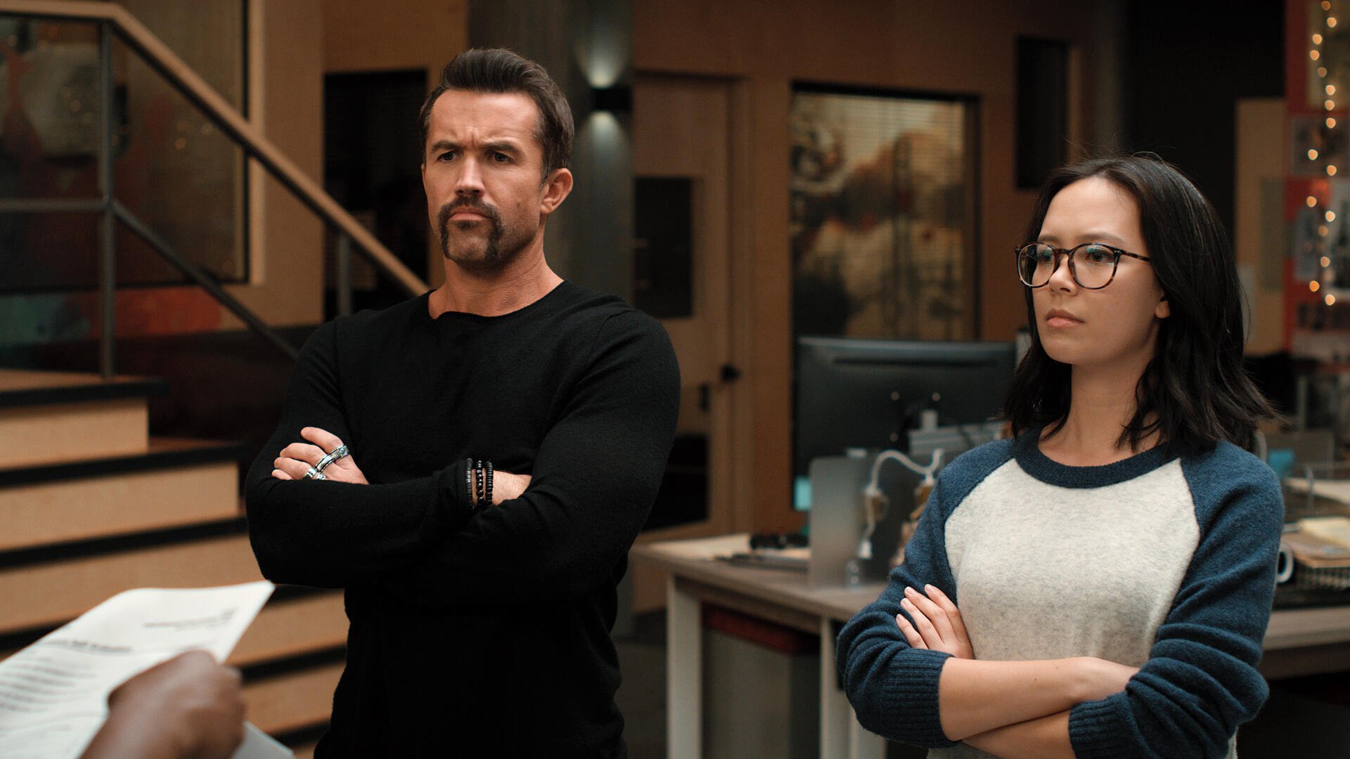 A scene from Mythic Quest featuring Rob McElhenney and Charlotte Nicdao, both with serious expressions, standing in a modern office environment. 4K Ultra HD wallpaper.