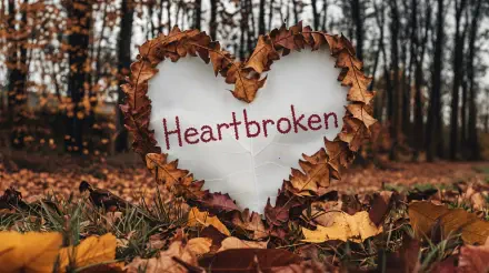 A heart-shaped arrangement of autumn leaves surrounds the word Heartbroken, set against a blurred forest background. This 4K Ultra HD wallpaper captures a poignant atmosphere.