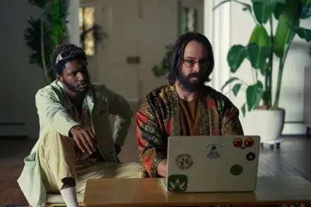 Two men are seated indoors, one working on a laptop while the other observes. The scene captures a moment from the TV show Tulsa King. 4K Ultra HD desktop wallpaper.