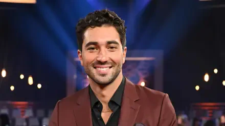 Joey Graziadei smiles confidently, dressed in a stylish suit against a backdrop of dramatic stage lighting, showcasing his role on Dancing with the Stars in this HD wallpaper.