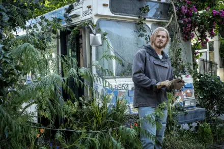 A scene from the TV show Earth Abides featuring Alexander Ludwig surrounded by lush greenery and an overgrown bus, creating an evocative post-apocalyptic atmosphere.