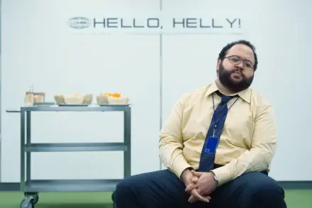 A scene from the TV show Severance featuring Zach Cherry, sitting in an office setting with a table of snacks and a sign reading HELLO, HELLY! in the background. 4K Ultra HD wallpaper.