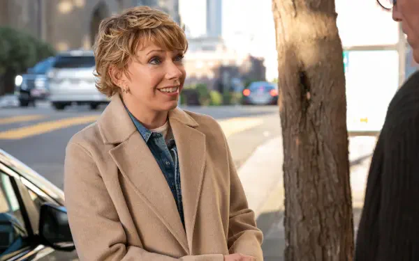 A scene from the TV show A Man on the Inside featuring Mary Elizabeth Ellis, smiling and engaging in conversation outdoors, framed by a tree and city backdrop.