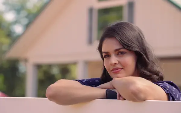 A woman leans on a white fence, gazing thoughtfully in front of a charming house, capturing a serene moment from the TV show Tulsa King, showcased as a 4K Ultra HD desktop wallpaper.