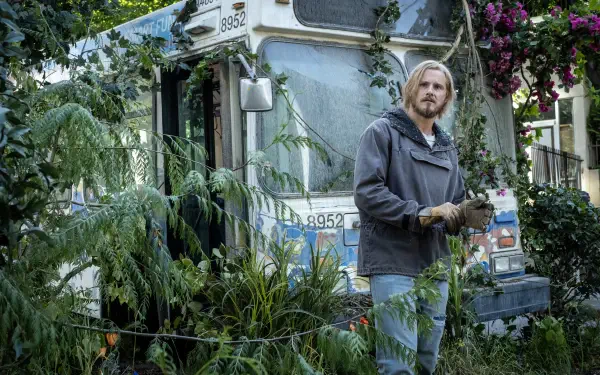 A scene from the TV show Earth Abides featuring Alexander Ludwig surrounded by lush greenery and an overgrown bus, creating an evocative post-apocalyptic atmosphere.