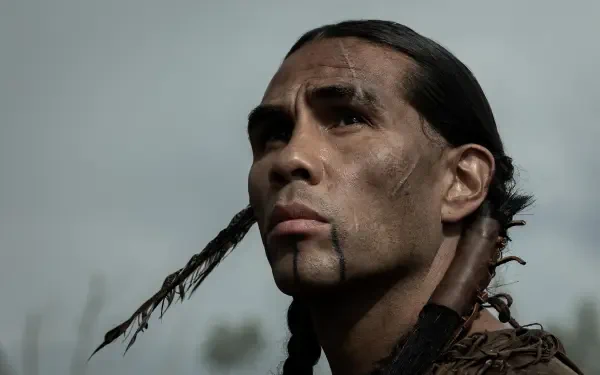 A dramatic HD wallpaper from the TV show American Primeval, featuring a focused character with long hair and facial markings, set against a moody, cloudy backdrop.