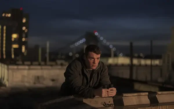 A dramatic scene from The Night Agent, featuring Gabriel Basso leaning against a rooftop barrier at night, with city lights and a bridge in the background. HD wallpaper-ready image.