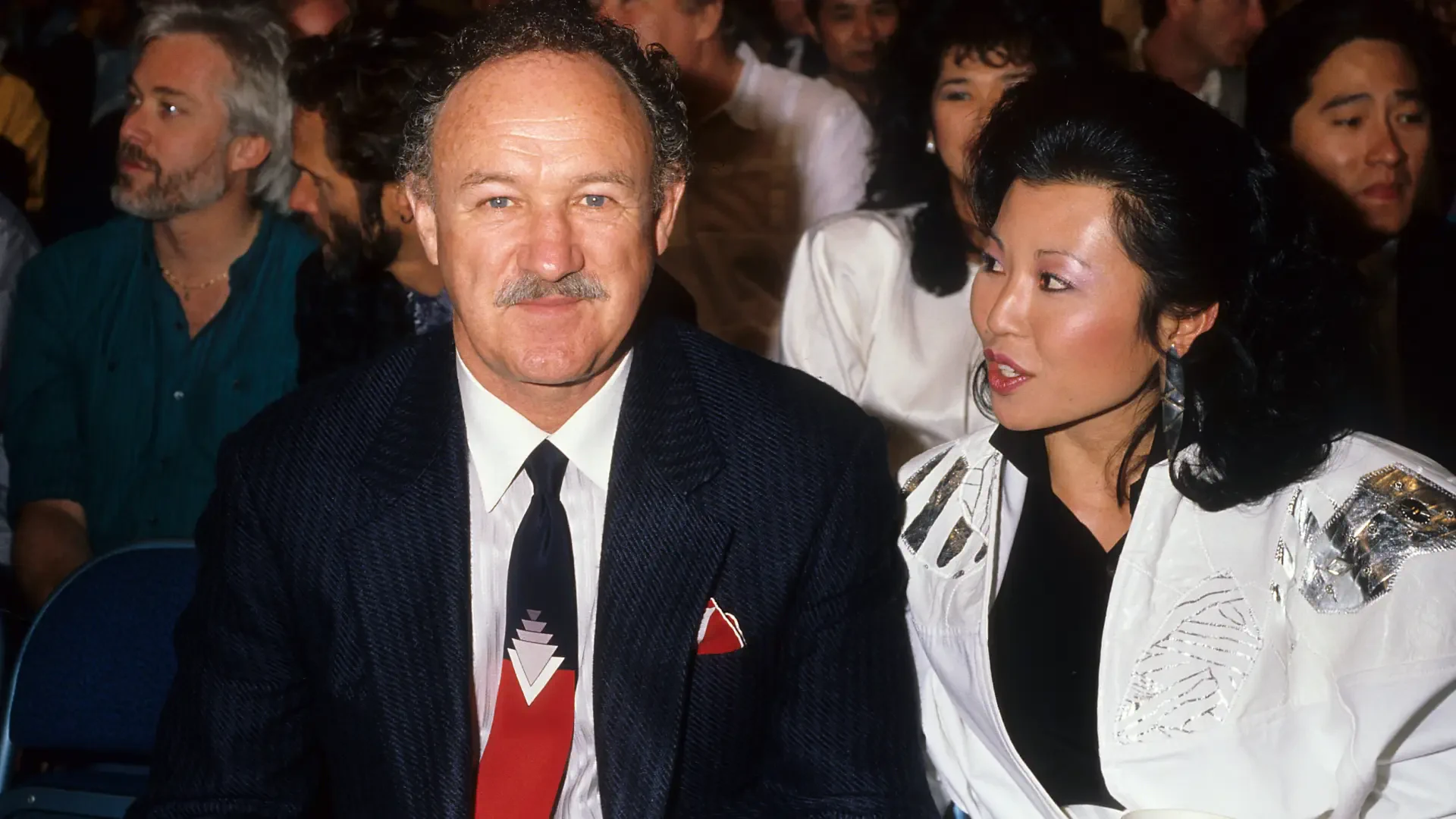 Gene Hackman seated with a companion at an event, captured in a candid moment. The background features a lively crowd, highlighting a vibrant atmosphere. HD desktop wallpaper.