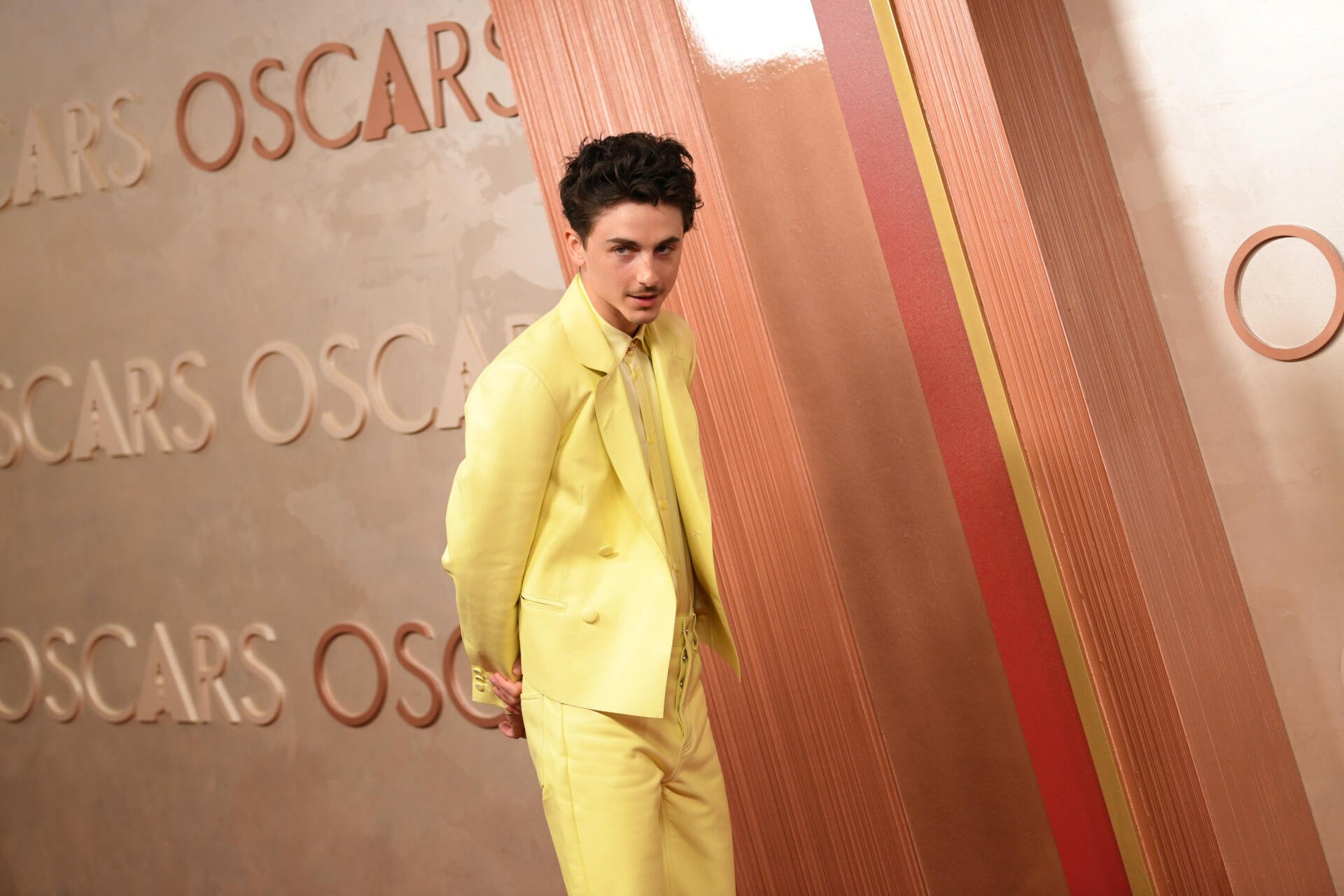 Timothée Chalamet poses in a striking yellow suit on the Oscars red carpet, against a backdrop featuring the iconic Oscars lettering. 4K Ultra HD Desktop Wallpaper.