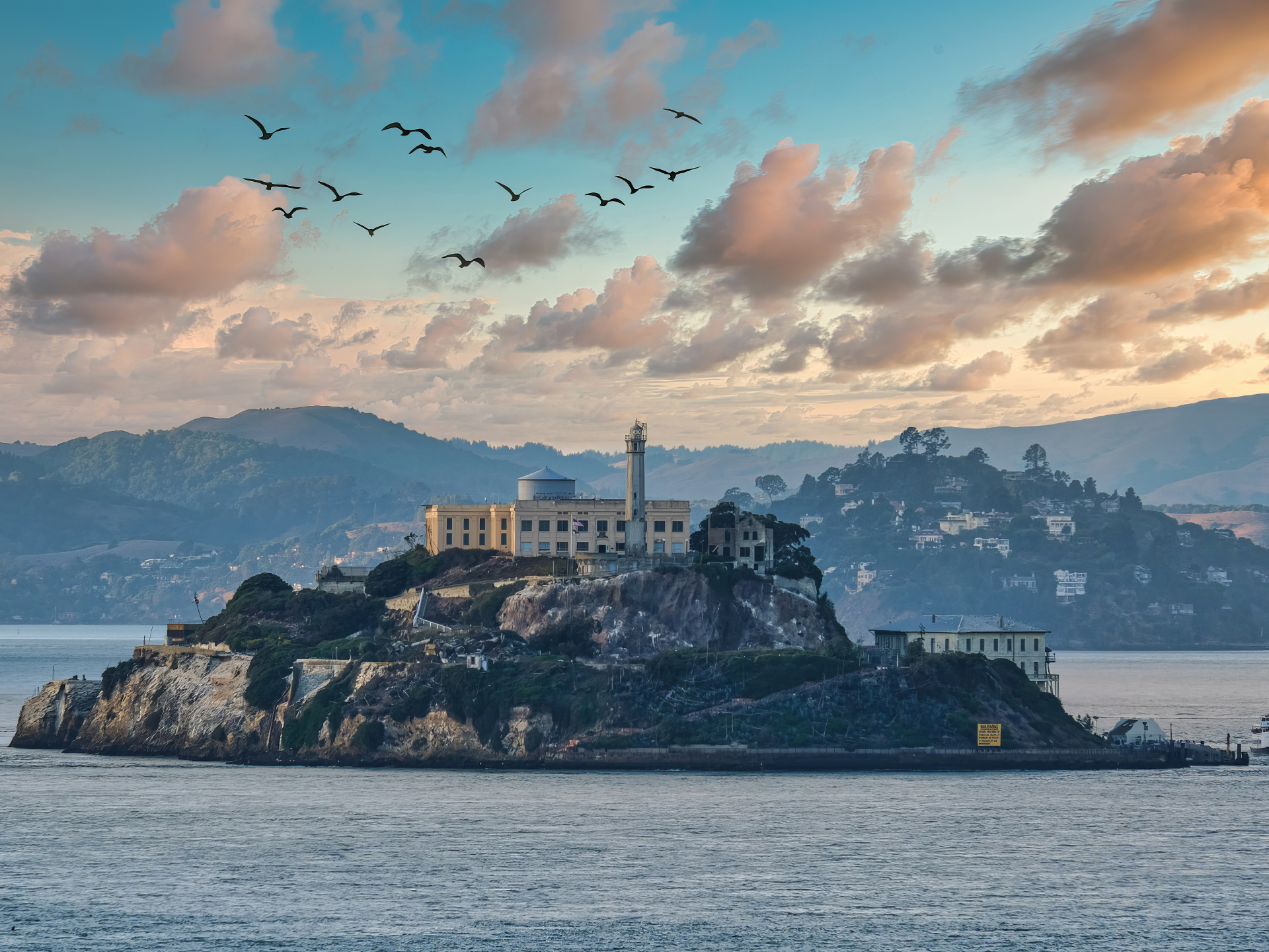 HD desktop wallpaper showing Alcatraz Island at sunrise with clouds, birds flying overhead, and the surrounding water and hills in the background.