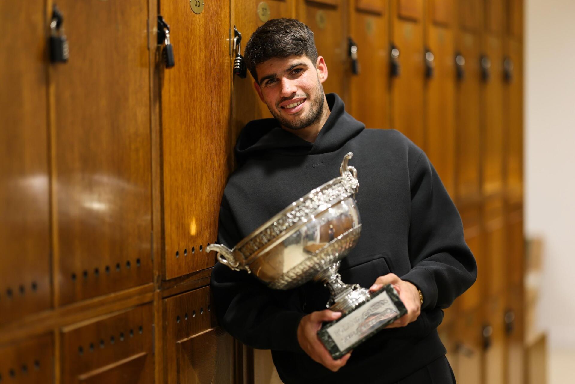 Carlos Alcaraz poses with a large trophy inside a wooden locker room, celebrating his victory at The French Open (Roland Garros) in this HD tennis-themed desktop wallpaper.