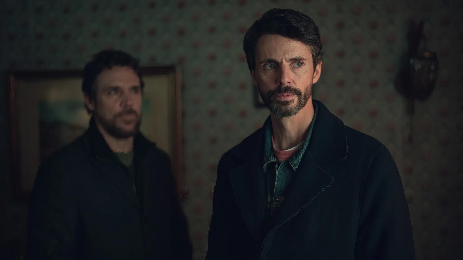 HD PC desktop wallpaper featuring Matthew Goode in the TV show Dept. Q, presenting a dark, suspenseful atmosphere with two serious characters in a dimly lit room.