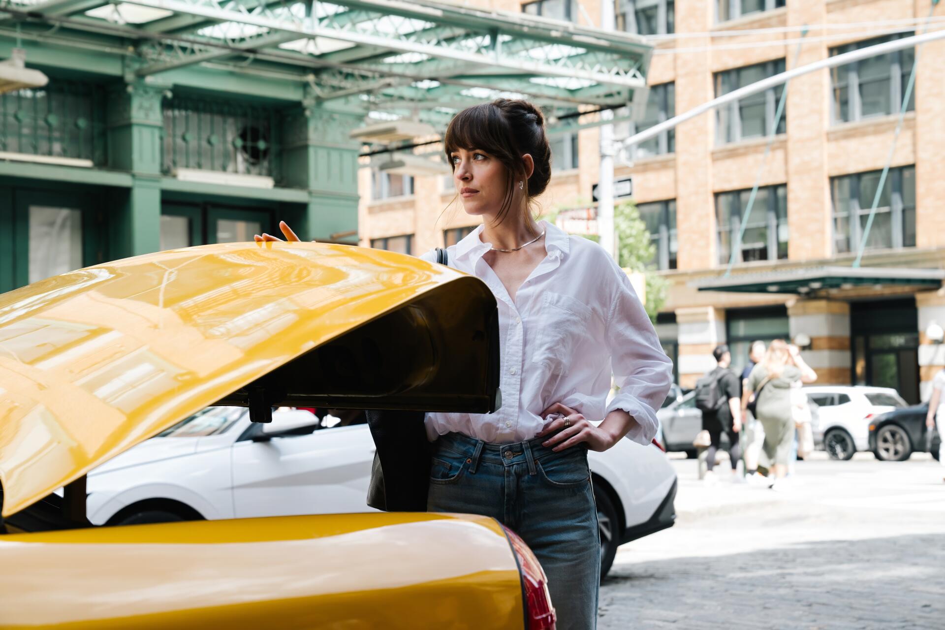 Dakota Johnson stands by an open yellow car trunk in an urban setting, featured in a 4K Ultra HD PC wallpaper from the movie Materialists.