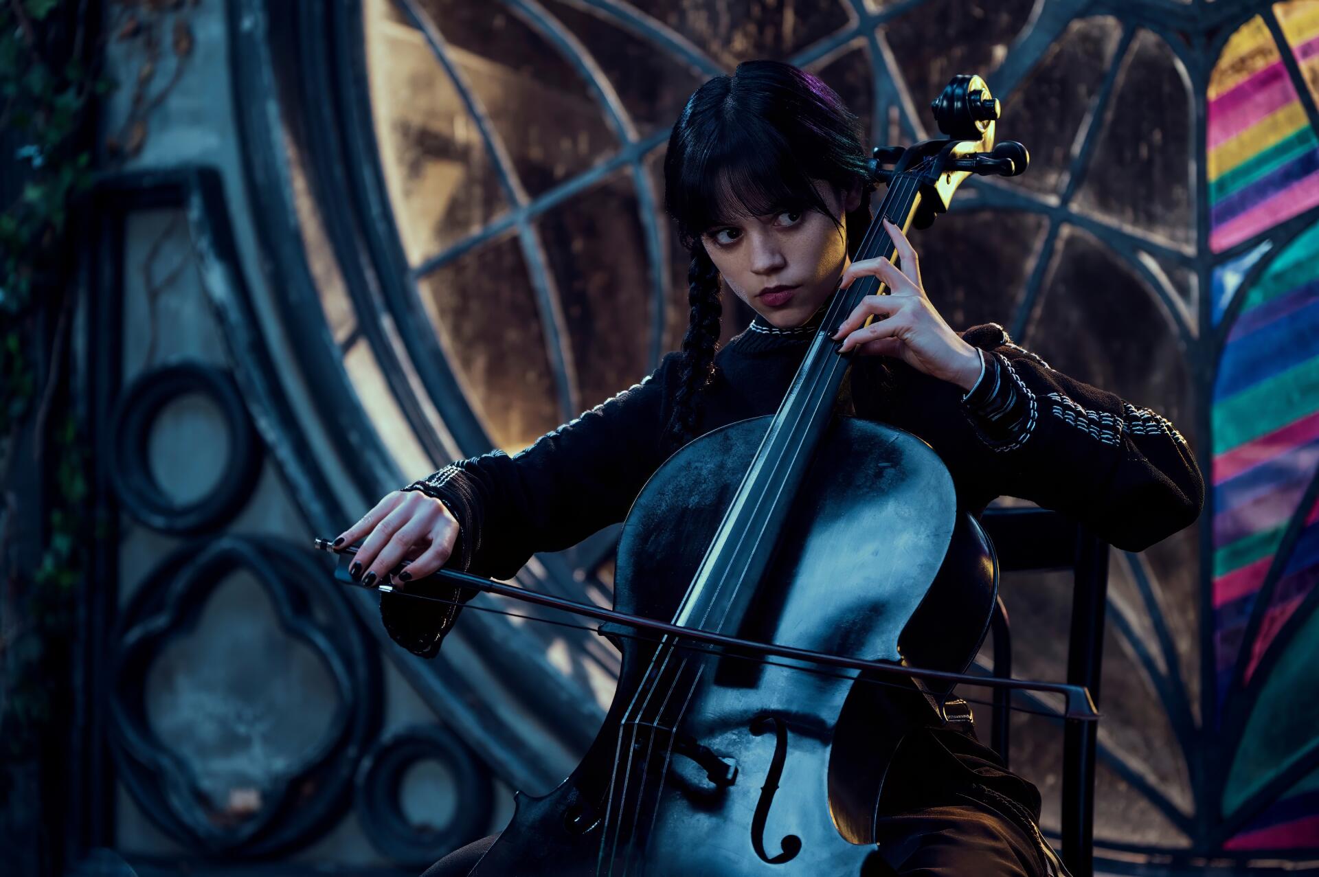 HD PC desktop wallpaper: moody scene from the TV show Wednesday — the lead character in gothic attire plays a cello against a dark, colorful, stained-glass backdrop.
