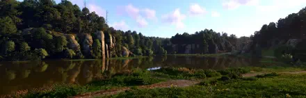 A serene lake surrounded by lush greenery and rocky cliffs, showcasing the vibrant nature found in Kingdom Come: Deliverance II. This HD image serves as a stunning desktop wallpaper.