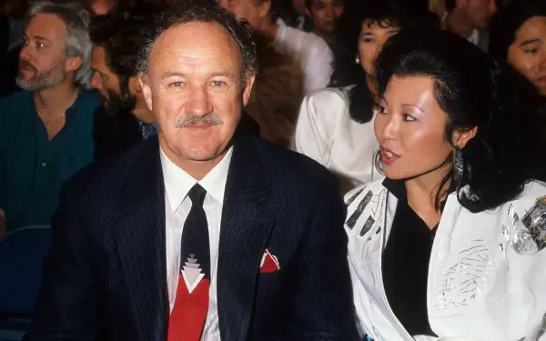 Gene Hackman seated with a companion at an event, captured in a candid moment. The background features a lively crowd, highlighting a vibrant atmosphere. HD desktop wallpaper.