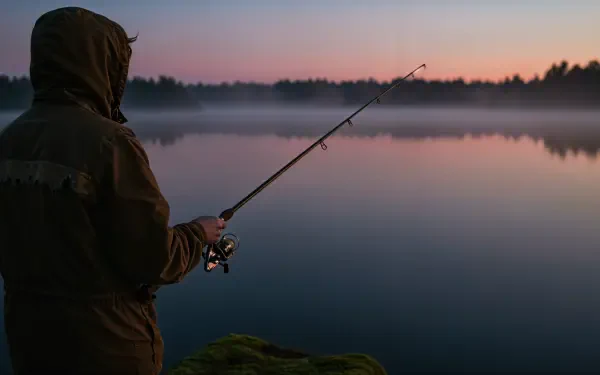 A person holding a fishing rod stands by a calm lake at dawn, surrounded by mist and trees, creating a serene atmosphere. This image serves as a stunning 4K Ultra HD wallpaper.