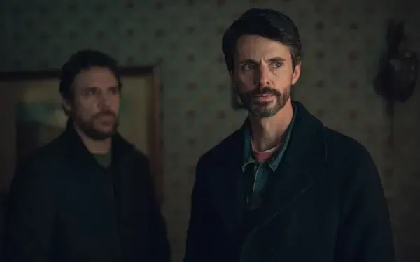 HD PC desktop wallpaper featuring Matthew Goode in the TV show Dept. Q, presenting a dark, suspenseful atmosphere with two serious characters in a dimly lit room.