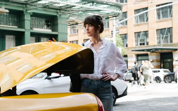 Dakota Johnson stands by an open yellow car trunk in an urban setting, featured in a 4K Ultra HD PC wallpaper from the movie Materialists.