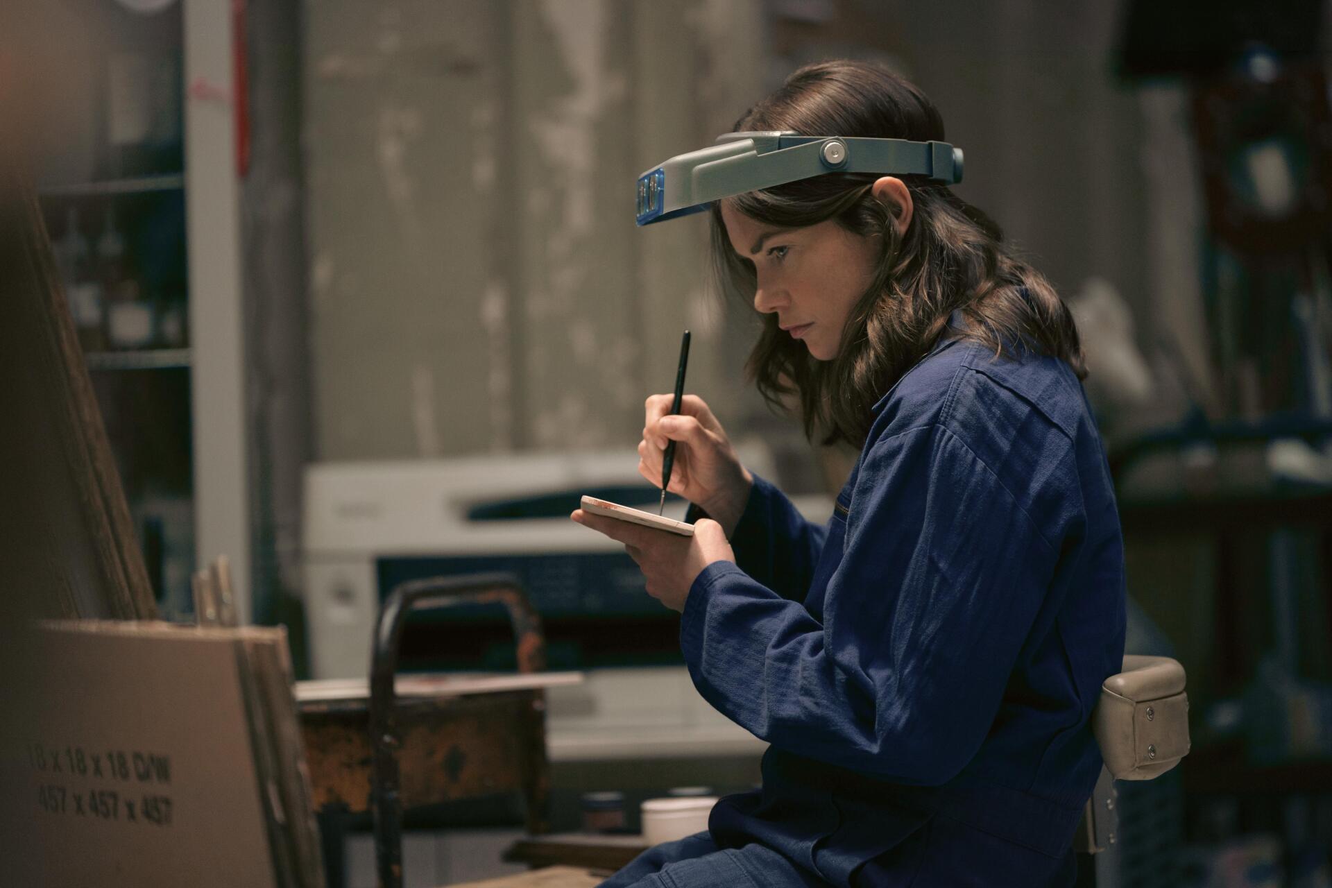 5K UHD PC desktop wallpaper of a woman in a dim workshop jotting notes, a moody scene from the TV show Down Cemetery Road.