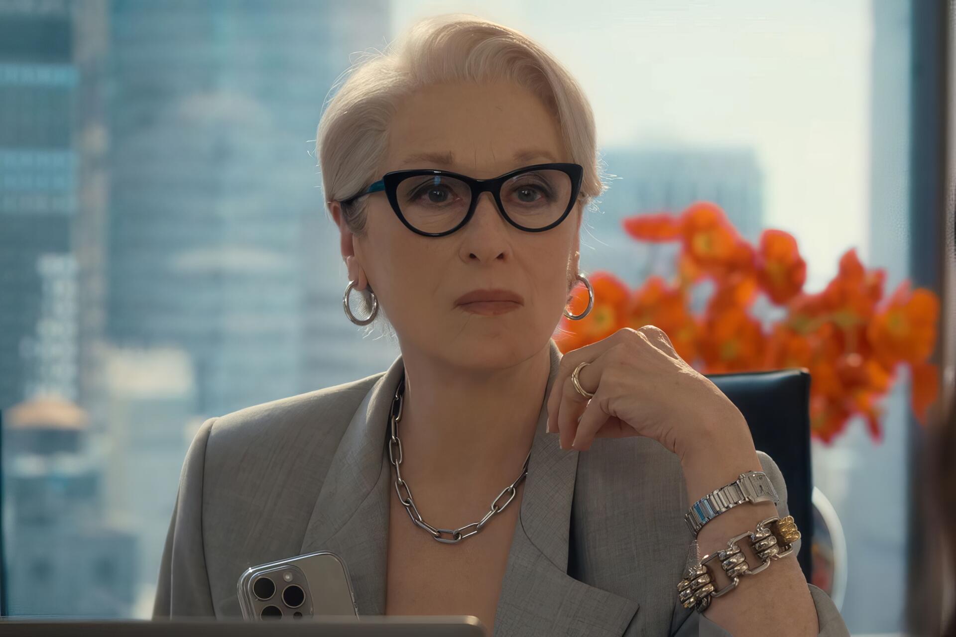 HD PC desktop wallpaper of a poised, silver‑haired actress in glasses and a gray suit from the movie The Devil Wears Prada 2, seated in a city office with orange flowers behind her.
