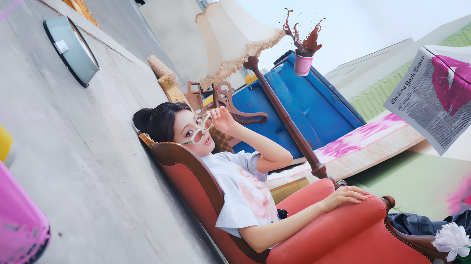 Woman wearing shades lounges in a red armchair amid colorful props, striking a fashion-model pose — vibrant 4K desktop wallpaper.