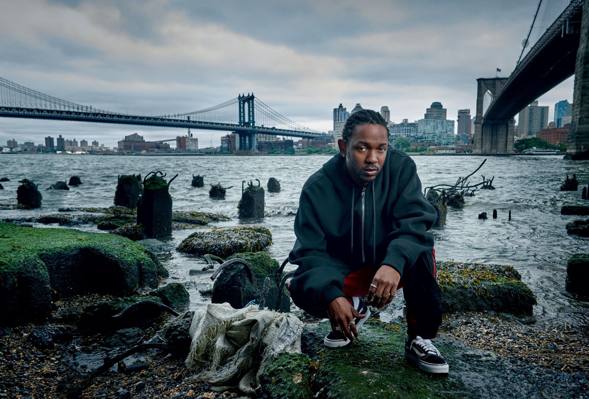 4K Ultra HD PC wallpaper: a hip-hop artist crouches on a rocky East River shore beneath the Manhattan Bridge in New York, moody urban skyline and textured waterfront background.