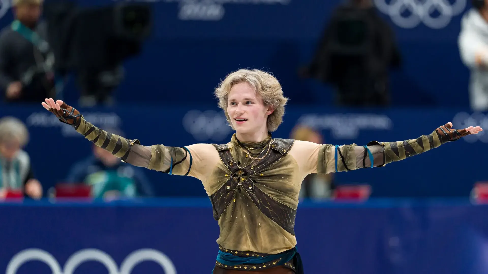 Ice skater in an ornate costume, arms outstretched on the Winter Olympics rink — dynamic sports action captured as a 2K Quad HD PC desktop wallpaper background.