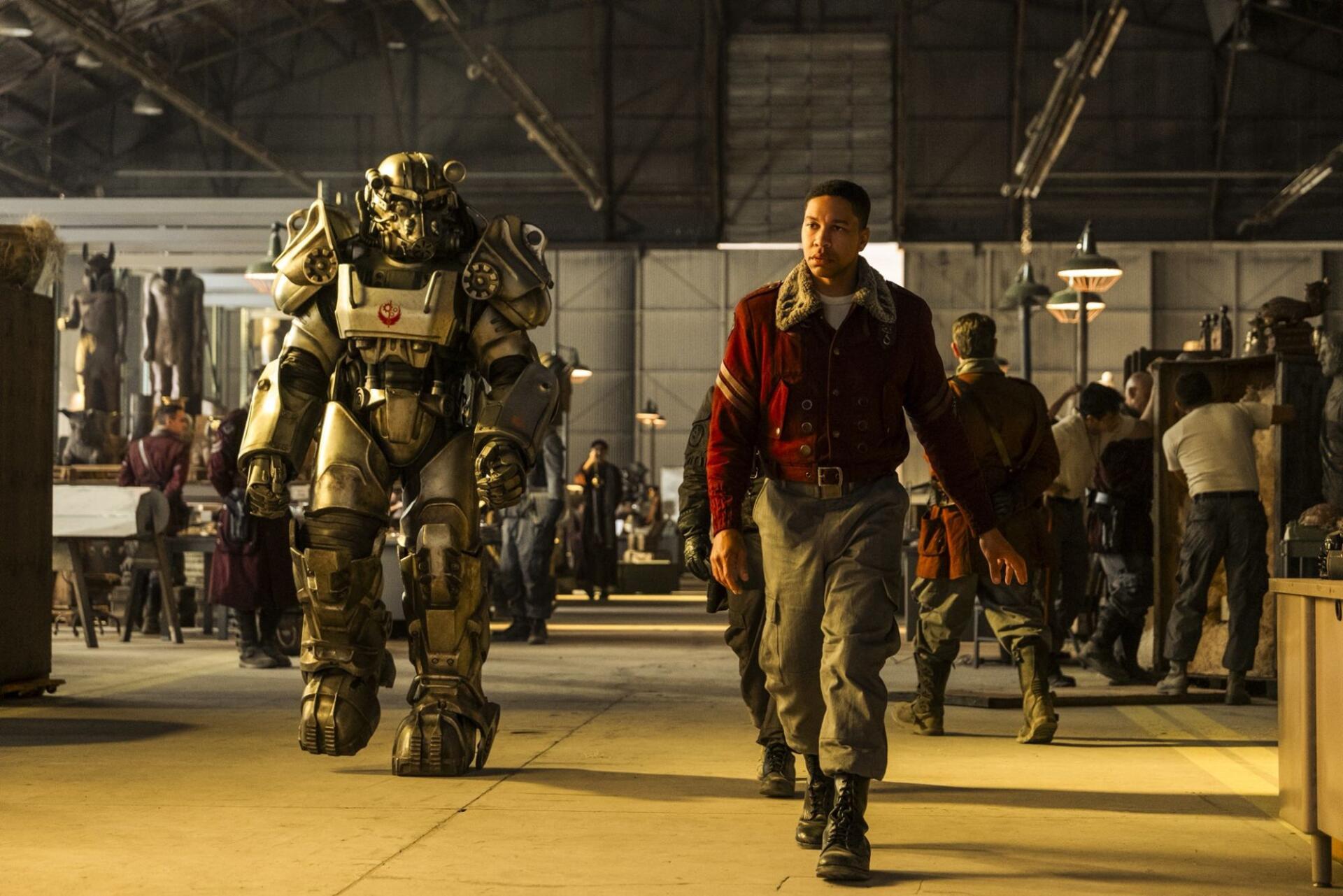 Man in a red coat strides through an industrial hangar beside a towering power armor suit, figures and gear in the background — Fallout (TV Show) HD PC desktop wallpaper and background.