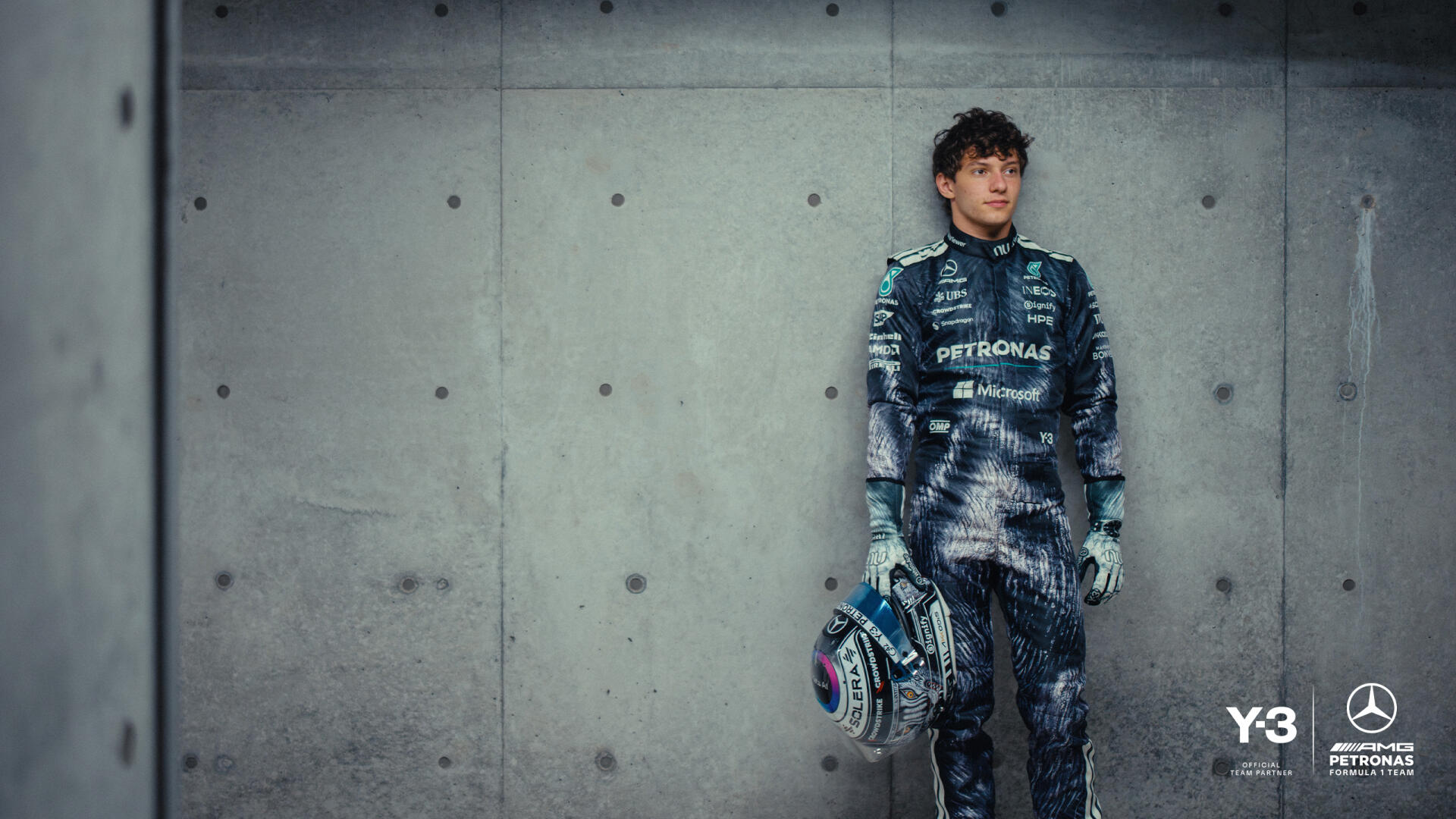 F1 driver in Mercedes-AMG race suit holding a helmet, leaning against a concrete wall — 2K Quad HD PC desktop wallpaper background