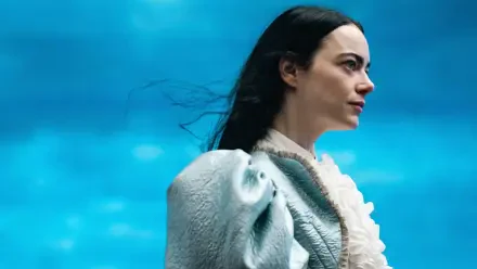 Woman in a pale ruffled period dress and cloak gazes right against a vivid blue sky — Poor Things 4K Ultra HD PC desktop wallpaper.