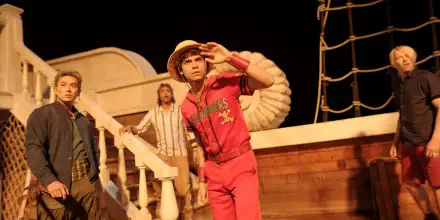 One Piece (Live Action) 2K Quad HD wallpaper: straw-hat captain in a red tracksuit scans the horizon aboard a wooden ship with two crewmates under warm stage lighting.